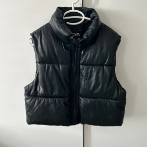 Svart dunväst från Zara - Snygg svart dunväst från Zara med quiltad design och hög krage. Västen har en grov dragkedja framtill och är tillverkad i ett skinnliknande syntetmaterial som ger en cool vibe. Perfekt att slänga över en hoodie eller tröja för extra edge. Pris 150kr, ordinarie 499kr. Lite defekt 3 bilden!