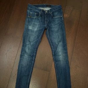Blå skinny jeans från Tiger of Sweden - Snygga blå jeans från Tiger of Sweden med skinny passform och klassisk femficksdesign. Jeansen har en mörk tvätt med slitningar och är tillverkade i stretchig denim för en bekväm känsla. Perfekta för dig som gillar en smal siluett. OBS! Har ett snus märke på ena bak fickan