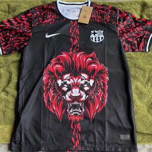 FC Barcelona x Nike svart lejontröja - Säljer en fet svart FC Barcelona x Nike fotbollströja i Dri-FIT-material. Tröjan har korta ärmar, röd och svart grafisk design med ett stort lejontryck på framsidan och klubbmärke på bröstet. Perfekt för dig som vill sticka ut på planen eller läktaren.