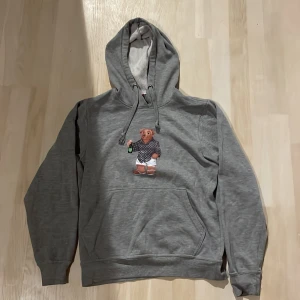 Grå hoodie med nalletryck - Grå hoodie med en stor luva och snörning. På bröstet finns ett tryck av en nallebjörn i skjorta och shorts som håller en flaska. Tröjan har magficka och ribbade muddar vid ärmslut och nederkant. Perfekt för en chill och avslappnad stil.