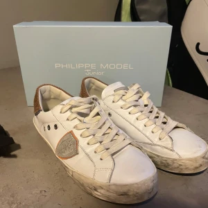 Philippe Model vita sneakers junior - Snygga vita sneakers från Philippe Model Junior med bruna detaljer i manchester baktill och grått sköldmärke på sidan. Skorna har snörning, platt sula och coola metallnitar vid snörhålen. Använd cirka 10 eller 12 gånger. Storlek 37 men passar typ 37 1/2 och kanske storlek 38. Låda ingår !