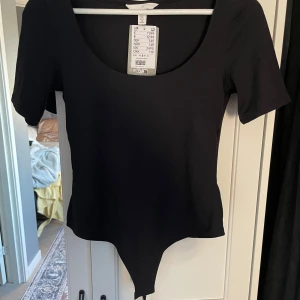 Svart kortärmad bodysuit från H&M - En svart, enkel och stilren bodysuit från H&M med korta ärmar och rundad urringning. Perfekt att styla med jeans eller kjol för en clean look.