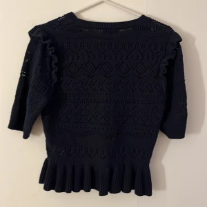 Svart stickad blus med volangdetaljer - Säljer en mörk blå  stickad blus med snyggt hålmönster och volanger längs axlarna. Från holly & whyte by lindex