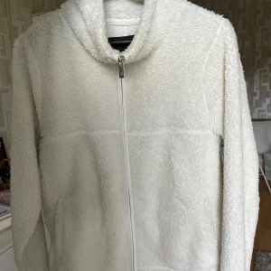 Vit teddyjacka med dragkedja - Mysig vit teddyjacka med huva och hel dragkedja framtill. Jackan har en fluffig och mjuk yta, perfekt för kyliga dagar. Långärmad modell med två fickor på sidorna. Enkel och stilren design som är lätt att matcha.