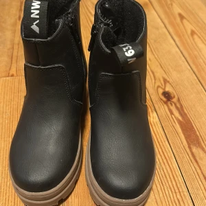 Svarta boots från H&M med grov sula - Svarta boots från H&M med grov, beige sula och dragkedja på sidan. Skorna är i syntetmaterial och har rund tå samt praktisk ögla baktill. Perfekta för höst och vinter när du vill ha en stilren look med extra grepp.