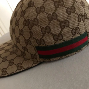 Gucci keps med GG-mönster - Säljer en beige keps från Gucci med klassiskt GG-monogram i brunt över hela kepsen. På sidan finns den ikoniska gröna och röda randen. Kepsen har böjd skärm och justerbar rem baktill. Tillverkad i Italien och i slitstarkt bomullsmaterial. Knappast använd har kvar Gucci lådan med kvitton kvar. 2500 i denna skick är sällsynt att få tag på!
