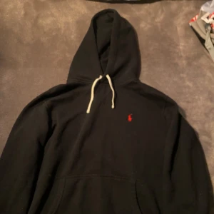 Svart hoodie från Polo Ralph Lauren - Svart hoodie från Polo Ralph Lauren med klassisk röd logga broderad på bröstet. Tröjan har huva med vita snören och en stor magficka framtill. 8/10 skick och inga defekter alls! Hör av er om frågor:)