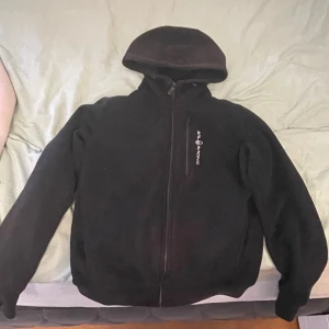 Svart fleecehoodie från Sail Racing XS - Mysig svart hoodie från Sail Racing i fleece med dragkedja framtill och en bröstficka med logga. Perfekt för kyliga dagar, har huva och långa ärmar. Enkel och stilren design som funkar till det mesta.