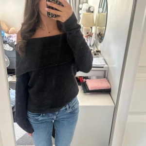 Svart offshoulder stickad tröja - Mysig svart stickad tröja med offshoulder-design och långa ärmar. Tröjan har en bred krage som vilar snyggt över axlarna och ger en avslappnad men trendig look. Perfekt att matcha med jeans för en cool vardagsstil.