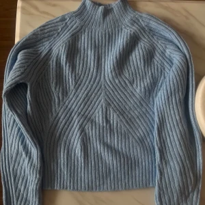 Ljusblå ribbad stickad tröja - Superfin ljusblå stickad tröja med ribbat mönster och hög hals. Tröjan har långa ärmar och en snygg struktur som ger en modern vibe. Perfekt att styla med jeans eller kjol för en chill look.