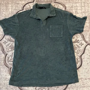 Grön frotté t-shirt från 157 - Mjuk grön t-shirt från 157 i frottématerial med korta ärmar, krage och en bröstficka. Perfekt för chill dagar och har en relaxed vibe. Snygg att matcha med jeans eller shorts för en laidback stil. Kan diskuteras!