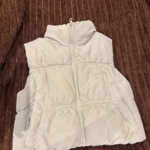 Vit dunväst i XS med hög krage - Säljer en vit dunväst i storlek XS med hög krage och dragkedja framtill. Västen har två fickor på sidorna och är quiltad för extra värme. Perfekt att slänga över en hoodie för en cool lager-på-lager-look. Den har en fläck vet inte om den går bort. Den är väldigt liten i storleken.