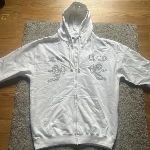 Vit hoodie från Bling Lucid med strass - Vit hoodie med dragkedja från Bling Lucid. Framsidan har coola strassdetaljer med texten 'BLING' och 'LUCID' samt två änglar. Tröjan har huva med snörning och är tillverkad i bomull och polyester. Perfekt för dig som gillar statement-plagg.