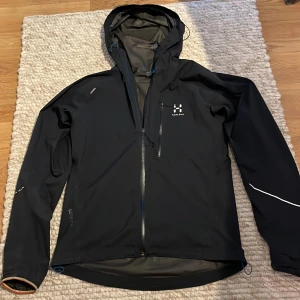 Svart vindjacka från Haglöfs med huva - Svart vindjacka från Haglöfs med huva och hel dragkedja framtill. Jackan har en bröstficka med dragkedja, diskreta reflexdetaljer på ärmarna och justerbar nederkant. Tillverkad i slitstark polyester med GORE-TEX för extra väderskydd, den kostar även 2800 ny pris.