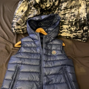  Moncler Väst - Säljer en blå quiltad dunväst från Moncler, modell Gilet L.Saison Patrick. Västen har en snygg huva, två dragkedjeförsedda fickor och ikonisk Moncler-logga på bröstet. Perfekt för lager-på-lager och streetstyle. Fodret är mjukt och materialet är glansigt.
