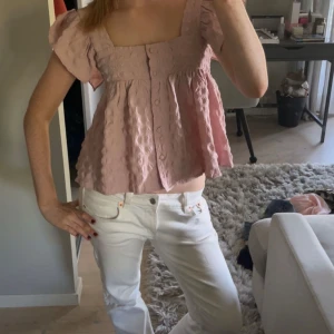 Ljusrosa blus med puffärm och knappar - Supersöt ljusrosa blus med puffiga korta ärmar och knappar framtill. Blusen har en fyrkantig urringning och ett luftigt, volangigt fall. 