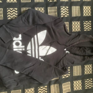 Svart Adidas hoodie - Svart hoodie från Adidas med den klassiska vita treklöver-loggan och stor Adidas-tryck på framsidan. Tröjan har huva med snörning och långa ärmar. Mjuk bomullskvalitet som är skön att ha på sig. Perfekt för chill eller streetwear-stil.