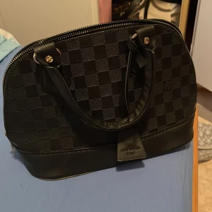 Svart handväska  - Snygg svart handväska  med klassiskt rutigt mönster och rundad form. Väskan har två handtag, detaljer i skinn och en dragkedja upptill. Perfekt för dig som gillar stilrena och lyxiga accessoarer.