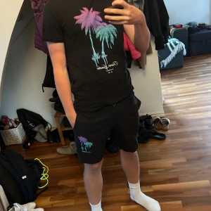 Svarta shorts+tröja Closure London med palmer - Svarta shorts+Tröja från Closure London med färgglada palmer i lila och turkos tryck på ena benet. Mjuk bomullskänsla, ribbad midja med snörning och normal passform. Perfekt för chill dagar och sommarhäng.