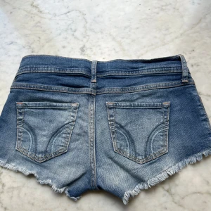Lågmidjade hollister shorts - Snygga blå jeansshorts från Hollister med fransig kant och klassiska fickor både fram och bak. Shortsen har låg midja och en slitning som ger en cool, avslappnad look. Perfekta för varma dagar.