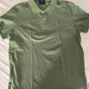 Grön pikétröja från GANT - Snygg grön pikétröja från GANT i klassisk modell med krage och knappar. Tröjan har korta ärmar och en liten broderad logga på bröstet. Perfekt för en stilren och avslappnad look.