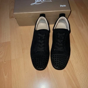 Svarta sneakers med nitar från Christian Louboutin - Säljer ett par svarta sneakers från Christian Louboutin med svarta nitar på tån och klassisk röd sula. Skorna har snörning och är tillverkade i skinn med en stilren och lyxig design. Kommer med originalkartong och dustbag. Skriv för mera info, priset är inte hugget i sten!😁