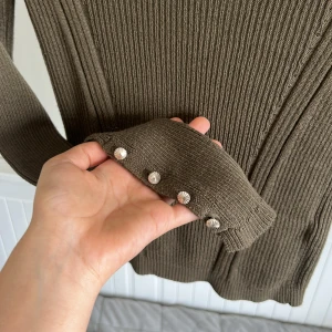 Olivgrön ribbad polotröja från Topshop - Säljer en olivgrön ribbad polotröja från Topshop. Tröjan har långa ärmar med dekorativa silverfärgade knappar vid ärmsluten och en figurnära passform. Perfekt att styla till jeans eller kjol! Priset går att diskutera 