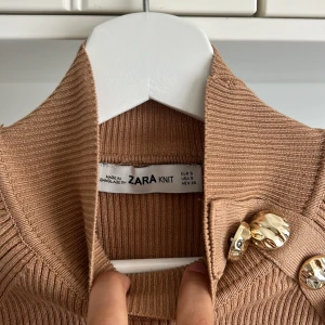 Beige ribbad stickad tröja från Zara med guldknappar - Säljer en beige ribbad stickad tröja från Zara med hög krage och dekorativa guldknappar längs axeln. Tröjan har lång ärm och en snygg, lite croppad passform. Perfekt för att lyfta din outfit med en elegant touch. Priset går att diskutera 