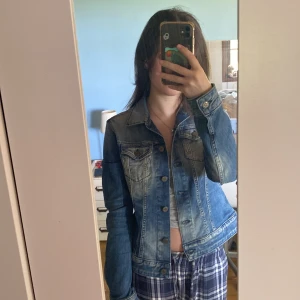 Blå jeansjacka från Replay - Säljer en klassisk blå jeansjacka från Replay med snygga slitningar och bronsfärgade knappar. Jackan har två bröstfickor med lock och knappar samt markerade sömmar för en cool look. Perfekt att slänga över en t-shirt eller hoodie.