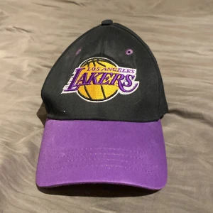 Los Angeles Lakers keps med broderad logga - Säljer en svart och lila keps med broderad Los Angeles Lakers-logga framtill och text baktill. Justerbart spänne i metall baktill. Perfekt för dig som gillar basket och vill visa ditt lagstöd.
