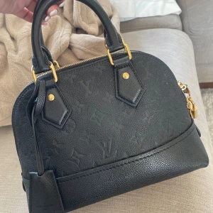 Svart handväska från Louis Vuitton - Säljer en svart handväska från Louis Vuitton med klassiskt monogrammönster i lädret. Väskan har guldfärgade metalldetaljer, dubbla handtag och dragkedja upptill. Perfekt storlek för det viktigaste och en lyxig accessoar till din outfit. Knappt använd. Den har dock en liten fläck som syns på sista bilden som troligen kan tas bort. Köpt på Louis Vuitton i Stockholm. Köpt för 18 900 kr. 💓