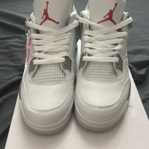 Nike Air Jordan 4 Retro “White Oreo” - Säljer ett par fräscha Nike Air Jordan 4 Retro i vitt med röda detaljer. Skorna har klassisk mesh på ovandelen, gråa partier vid sulan och ikonisk Jumpman-logga på plös och häl. Perfekta sneakers för dig som gillar streetwear.