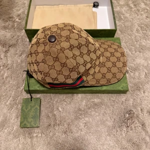 Beige keps från Gucci med GG-mönster - Säljer en beige keps från Gucci med klassiskt GG-monogram över hela kepsen. Kepsen har gröna och röda ränder på sidan och är tillverkad i bomullsmaterial. Kommer med originalask och dustbag. Denna keps är perfekt nu till sommaren!