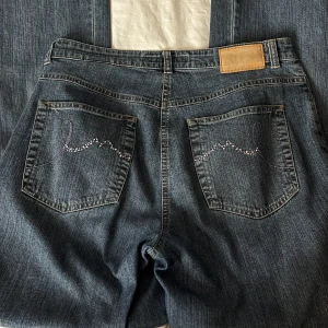 Blå jeansbyxor från MAC - Klassiska blå jeansbyxor från MAC med raka ben och normal passform. Byxorna har broderade detaljer på bakfickorna och stängs med knapp och dragkedja. Perfekta för unik stil.