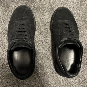 Svarta sneakers från Axel Arigato i mocka - Säljer ett par svarta sneakers från Axel Arigato i mocka med diskret guldfärgad logga på sidan. Skorna har snörning, rund tå och platt sula. Perfekta för en stilren och clean look.