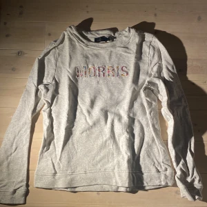 Grå sweatshirt från Morris - Säljer en ljusgrå sweatshirt från Morris med broderad logga på bröstet och en liten brodyr på ärmen. Tröjan har rund hals och långa ärmar. Tillverkad i mjukt bomullsmaterial.
