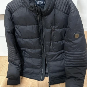 Svart pufferjacka från Jofama - Säljer en svart pufferjacka från Jofama med quiltade detaljer på axlar och ärmar. Jackan har dragkedja framtill och en bröstficka med dragkedja. Perfekt för kyligare dagar. Storlek M/L endast använd 4 gånger 