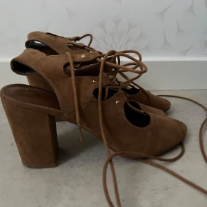 Bruna mockasandaler med klack och snörning - Säljer ett par bruna sandaler i mocka med hög klack och öppen tå. Skorna har snygg snörning framtill som går att knyta runt vristen för en cool look. Perfekta för att lyfta en outfit!