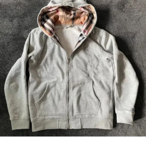 Grå hoodie med dragkedja från Burberry - Säljer en grå hoodie från Burberry med klassiskt rutigt foder i huvan och dragkedja framtill. Tröjan har två fickor och broderad logga på bröstet. Perfekt för en stilren och avslappnad look. Ända defekten, r dragkedjan som inte funkar att ta upp/: , där av priset!