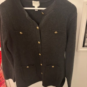 Mörkgrå kofta med guldknappar - Mörkgrå kofta med långa ärmar och fyra fickor framtill. Stängs med dekorativa guldknappar och har en klassisk, rak passform. Perfekt att slänga över en topp för en stilren look.