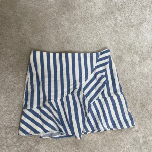 Blåvit randig kjol från Mango - Säljer en kort kjol från MNG med blå och vita ränder. Kjolen har omlott-design och är tillverkad i ett lätt material. 