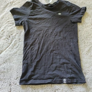 Svart t-shirt från DMG - Säljer en svart t-shirt från DMG med rund halsringning och diskret logga på bröstet samt nere vid fållen. T-shirten är enkel och stilren, perfekt till vardags. Tillverkad i mjukt bomullsmaterial.