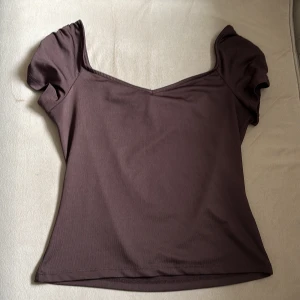 Brun topp med volangärmar - Säljer en brun topp med korta volangärmar och v-ringad hals. Toppen har en figurnära passform och är perfekt för att lyfta en enkel outfit. Superfin att styla med jeans eller kjol!