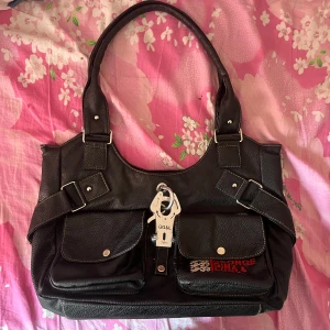 Svart handväska från George Gina & Lucy - Säljer en svart handväska från George Gina & Lucy med två framfickor, silvriga metalldetaljer och broderad logga i rött. Väskan har dubbla handtag och är tillverkad i syntetmaterial med struktur. Invändigt finns flera fack för organisering.