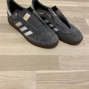 Adidas spezial i grå mocka - Grå Adidas Spezial med ovandel i mocka och klassiska vita ränder på sidorna. Skorna har brun sula och rund tå. Perfekta för dig som gillar retrostil och streetwear.