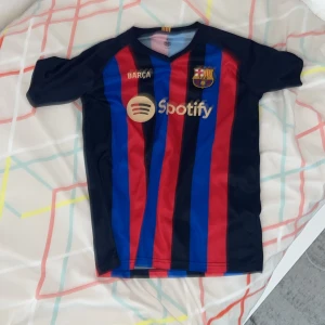 FC Barcelona matchtröja Pedri 8 - Säljer en FC Barcelona fotbollströja med Pedri 8 på ryggen. Tröjan har svarta, röda och blå vertikala ränder, korta ärmar och klubbmärke samt sponsorlogga på bröstet. Perfekt för dig som älskar Barça och vill visa ditt stöd för laget.