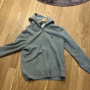 Grå hoodie från Polo Ralph Lauren -  Storlek: 14-16 år, liten i sig.   Säljer en klassisk grå hoodie från Polo Ralph Lauren med dragkedja och huva. Tröjan har två fickor framtill och den ikoniska lilla loggan broderad på bröstet. Perfekt för en avslappnad stil.