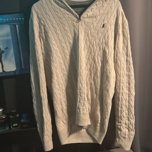 Vit stickad tröja med dragkedja från Polo Ralph Lauren - Säljer en vit stickad tröja från Polo Ralph Lauren med halv dragkedja och klassisk kabelstickad design. Tröjan har lång ärm och den ikoniska loggan broderad på bröstet. Perfekt för lager-på-lager och stilrena outfits.