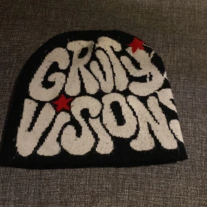 Svart och vit mössa med tryck - Cool svart mössa med vit text 'GRVTY VISIONS' och röda stjärnor på framsidan. Baksidan har ett stort vitt spindelnätsmönster. Perfekt för dig som gillar unika accessoarer med lite edge.