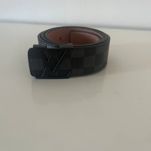 Svart rutigt bälte från Louis Vuitton - Säljer ett svart bälte från Louis Vuitton med klassiskt rutigt mönster och svart LV-spänne. Bältet har en brun insida och är tillverkat i skinn. Perfekt accessoar för att lyfta din outfit.